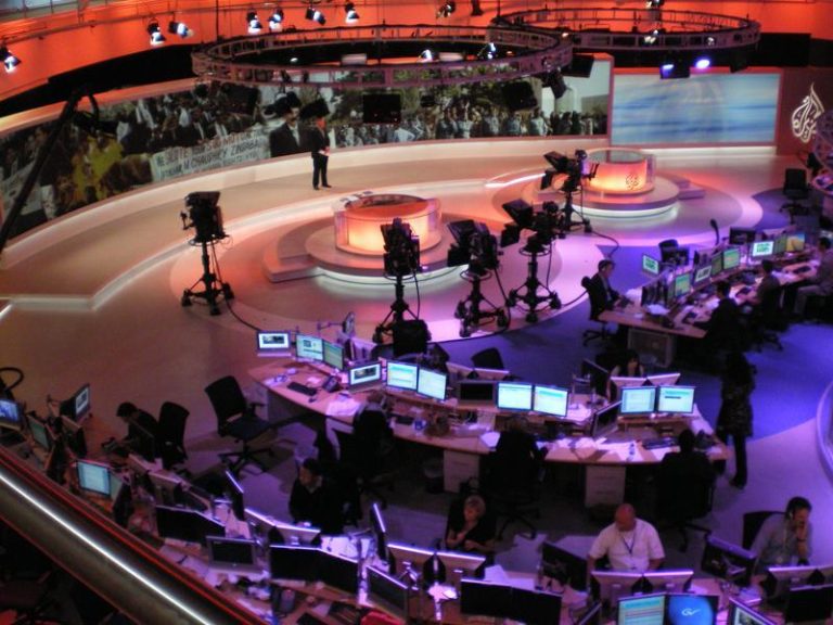 Journalists working at desks inside a busy television newsroom.